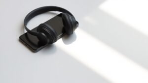 Modern wireless headphones positioned next to smartphone on clean off-white studio background. Soft side lighting creates subtle shadows. Both devices matte black finish. Clean minimal composition showing wireless connection concept. 16:9 horizontal format photorealistic product photography.