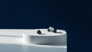 Pair of black wireless sport earbuds sitting on clean white surface with subtle shadows. Modern minimal product photography with soft side lighting. Deep navy blue background. Professional studio setup. No branding visible.