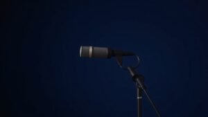 Professional condenser microphone mounted on boom stand against deep navy studio background with soft side lighting creating gentle shadows. Clean minimal composition showing the microphone capsule and shock mount detail in sharp focus with shallow depth of field.