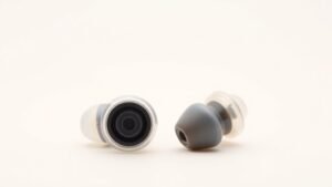 Close-up product photography of two in-ear monitors side by side on clean off-white background. One transparent shell showing single driver, one showing multiple drivers inside. Soft side lighting creates subtle shadows. Modern minimal composition horizontal format.