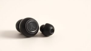 Close-up product shot of premium true wireless earbuds on clean off-white studio background. One earbud positioned to show inner sensor area. Soft directional lighting reveals subtle detail and technology components. Modern minimal composition with shallow depth of field.