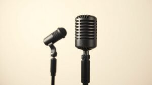 Two professional condenser microphones positioned at different angles on boom stands against a clean off-white studio background. Soft directional lighting from the left creates subtle shadows. The microphones show contrasting polar pattern diagrams subtly visible on their grilles.