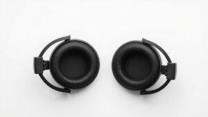 Professional overhead view of open-back headphones on clean off-white studio surface showing driver construction detail. Soft side lighting highlights acoustic elements and frequency response characteristics. Modern minimal product photography style.