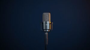 Professional condenser microphone on boom stand against deep navy studio background. Warm side lighting creates subtle highlights on metal grille and body. Clean minimal composition with soft shadows.