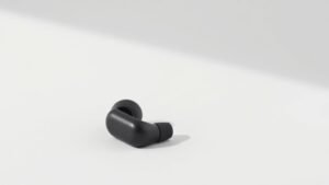 A single pair of sleek wireless sport earbuds resting on a minimalist off-white studio surface, shot from a low three-quarter angle. The earbuds feature a compact streamlined silhouette with rubberized ear fins and a matte finish. Soft directional side lighting casts subtle shadows that emphasize the contours and texture of the product. Clean negative space surrounds the subject. Photorealistic, 16 by 9 horizontal composition.