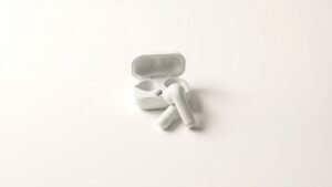 A single pair of compact wireless earbuds resting on a smooth off-white studio surface, the earbuds positioned neatly side by side with their small charging case open behind them. Soft directional side lighting casts gentle shadows that define the earbuds sleek contours and glossy finish. The background fades to a clean, seamless off-white tone, giving the composition a minimal, contemporary feel.