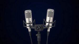 A matched pair of small diaphragm condenser microphones mounted in an XY stereo configuration on a professional shock mount, positioned against a deep navy background with soft directional side lighting. The microphones are silver and black, photographed from a slight angle, photorealistic, 16 to 9 horizontal format, no people, no text.