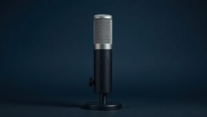 A single large-diaphragm condenser microphone stands upright on a sleek black desktop microphone stand, centered against a deep navy studio background. Soft diffused side lighting casts gentle shadows that highlight the metallic mesh grille and cylindrical body of the microphone, emphasizing its professional build quality and elegant industrial design. The composition is clean, minimal, and sharply focused with rich tonal depth.