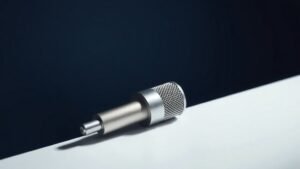 A sleek compact lightning connector microphone rests on a minimal white surface against a deep navy background. Soft directional studio lighting falls from the left, casting a gentle shadow and highlighting the brushed metal capsule and slim body. The microphone sits slightly angled, showcasing its compact form and polished connector tip with quiet confidence.