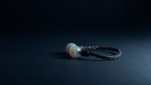 A single pair of in-ear monitors rests on a smooth matte surface against a deep navy studio background. The earpieces feature translucent shells with subtle internal detail visible beneath the surface. A braided cable coils naturally beside them. Soft directional side lighting catches the curved edges, casting a gentle shadow that adds depth and dimension to the minimal, elegant composition.