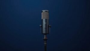 A single sleek condenser microphone stands upright on a minimal desktop mic stand, positioned slightly off-center against a clean deep navy background. Soft diffused side lighting sculpts the metallic body of the microphone, highlighting its cylindrical mesh grille and matte black finish. The composition is calm and editorial, with gentle shadows adding depth to the otherwise minimal studio scene.