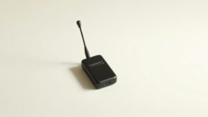 A single wireless lavalier microphone system displayed on a smooth off-white studio surface, the compact clip-on capsule resting beside its small bodypack transmitter unit. Soft directional side lighting casts gentle shadows that emphasize the sleek contours and subtle texture of the matte black finish. Clean negative space surrounds the arrangement, creating an elegant minimal composition with shallow depth of field.