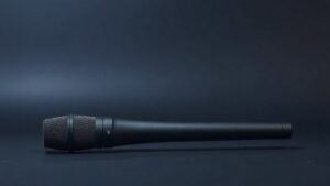 A single sleek shotgun microphone rests on a minimal studio surface, its long cylindrical barrel extending horizontally across the frame. The microphone features a matte black finish with subtle mesh detailing along the interference tube. Soft directional side lighting casts gentle shadows that emphasize the cylindrical form. The background is a smooth deep navy gradient, clean and uncluttered, giving the composition a modern professional feel.