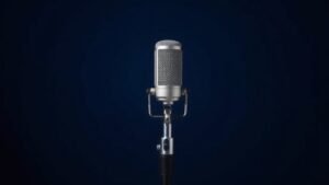 A single professional condenser microphone with a large diaphragm capsule stands upright on a sturdy desk stand, positioned slightly off-center against a clean deep navy background. Soft diffused side lighting catches the metallic mesh grille and polished chrome body, casting a gentle shadow to one side. The scene feels refined, minimal, and authoritative, suited to a serious audio review context.