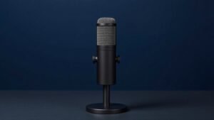 A single professional podcasting microphone stands upright on a sleek black desktop stand against a deep navy background. The microphone features a large diaphragm capsule with a fine metal mesh grille and a matte black cylindrical body. Soft diffused side lighting catches the subtle texture of the metal surface, casting a gentle shadow behind it. The composition is clean, minimal, and elegantly centered.