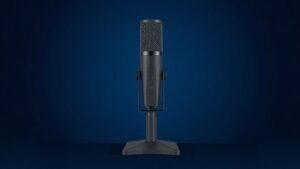 A single sleek USB condenser gaming microphone stands upright on a compact desktop stand, centered against a clean deep navy background. Soft directional studio lighting from the left catches the polished metallic mesh grille and matte black body, casting a subtle gradient shadow to the right. The composition is minimal and sharp, with rich tonal contrast highlighting the microphones modern angular form.