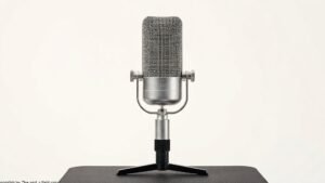A single professional condenser microphone stands upright on a sleek black desktop stand against a clean off-white studio background. Soft side lighting catches the metallic mesh grille and polished chrome body, casting a gentle shadow that adds depth. The microphone is centered with generous negative space, giving the composition a refined, editorial quality suited to a premium audio publication.