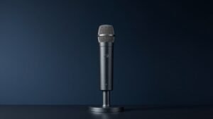 A single dynamic vocal microphone standing upright on a sleek matte black microphone stand against a deep navy studio background. Soft diffused side lighting catches the metallic mesh grille and the smooth cylindrical body, casting a gentle shadow to one side. The surface textures are crisp and detailed, giving the scene a clean, professional product photography feel.