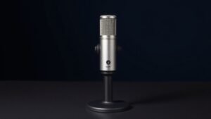 A single professional XLR microphone stands upright on a sleek black desk stand, positioned slightly off-center against a deep navy background. Soft side lighting from the left catches the brushed metal body, highlighting its cylindrical form and mesh grille with subtle metallic sheen. The composition is clean and minimal, with a gentle shadow falling behind the microphone on the smooth studio surface.