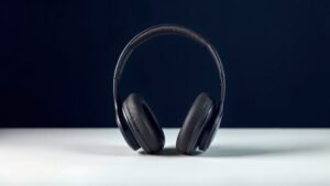 A single pair of premium over-ear headphones resting upright on a minimal white surface against a deep navy background. Soft diffused side lighting catches the smooth matte and polished surfaces, highlighting the cushioned ear cups and sleek headband arc. The composition is clean and centered with gentle shadows grounding the product. Studio quality, photorealistic, sophisticated and modern.