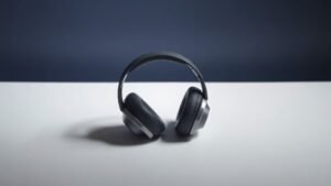 A single pair of sleek over-ear headphones resting on a minimal white surface against a deep navy background. Soft directional studio lighting from the left casts gentle shadows that define the cushioned ear cups and polished headband arc. The headphones face slightly to the right, showcasing their clean sculptural form with a premium matte and metallic finish. No clutter, no props.