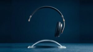 A single pair of premium over-ear audiophile headphones resting on a minimalist curved acrylic stand, shot against a deep navy studio background. Soft directional side lighting catches the brushed metal headband and cushioned leather ear cups, casting a gentle shadow across the surface below. The composition is centered, balanced, and refined, conveying high-end audio craftsmanship through clean modern product photography.