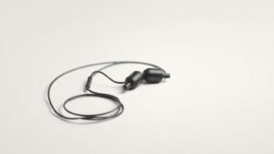 A single pair of in-ear monitors rests on a smooth off-white studio surface, their sleek earpieces angled slightly to reveal sculpted contours and detachable cables coiled neatly beside them. Soft directional side lighting casts gentle shadows that define the form and texture of the housings. The composition is minimal and precise, emphasizing refined industrial design against a clean, uncluttered background.