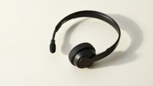 A single sleek headset microphone rests on a clean off-white studio surface, its slender boom arm curving gracefully toward a small capsule head. Soft side lighting casts gentle shadows that define the matte black finish and metallic accents. The minimalist composition emphasizes the lightweight headband and adjustable fit, conveying professional audio quality in a calm, uncluttered environment.