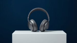 A single pair of sleek over-ear wireless headphones resting upright on a minimal white acrylic surface against a deep navy background. Soft diffused side lighting catches the smooth matte curves of the ear cups and polished headband, casting a gentle shadow beneath. The composition is clean and centered, emphasizing premium build quality and elegant modern design.