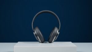 A single pair of sleek over-ear headphones resting upright on a minimal white acrylic surface against a deep navy background. Soft diffused side lighting catches the smooth matte ear cups and polished headband arc, casting a gentle shadow to one side. The composition is clean and centered with subtle depth, giving the scene a refined editorial studio quality.