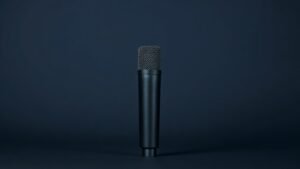 A single dynamic microphone stands upright on a minimal studio surface against a deep navy background. Soft side lighting catches the sleek cylindrical body, highlighting brushed metal mesh grille details and a matte black body with subtle sheen. The composition is clean and centered, with gentle shadow fall creating depth and dimension in a refined, editorial product photography style.