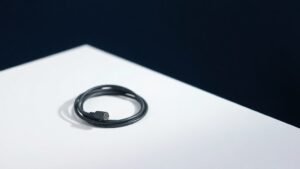 A single lavalier microphone with a slim cylindrical capsule and thin black cable coiled neatly beside it, resting on a smooth matte white surface against a deep navy background. Soft diffused side lighting catches the metallic sheen of the microphone head, casting a gentle shadow that adds depth and dimension to the minimal studio composition.