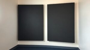 A pair of thick corner-mounted acoustic bass trap panels in a small professional studio, deep charcoal grey foam against an off-white wall, soft side lighting revealing texture and depth, clean minimal studio environment, no gear visible, photorealistic, 16 by 9 horizontal format, deep navy floor visible at base