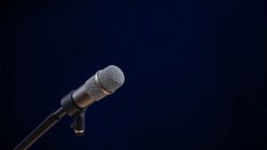 A single professional dynamic microphone mounted on a sleek desktop boom arm, angled slightly toward camera, centered against a deep navy studio backdrop. Soft diffused side lighting highlights the cylindrical body and fine mesh grille, casting a gentle shadow across the surface. The setup feels polished and broadcast-ready, with clean lines and a professional studio atmosphere.