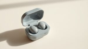 A pair of true wireless earbuds resting inside their open charging case, positioned at a slight angle on a smooth off-white surface. The case lid is propped open revealing the earbuds nestled inside. Soft directional studio lighting from the left casts gentle shadows, highlighting the glossy and matte surfaces of the compact earbuds. Clean minimal composition with shallow depth of field.