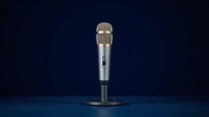 A single sleek USB condenser microphone stands upright on a minimal desk mount against a clean deep navy background. Soft diffused side lighting catches the brushed metal body and mesh grille, casting a subtle gradient shadow behind it. The microphone is centered in the frame with generous negative space, giving the composition a premium editorial feel.