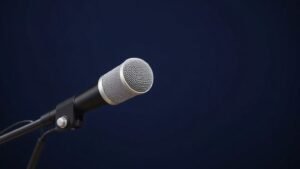A single large-diaphragm dynamic broadcast microphone mounted on a sturdy desktop boom arm, positioned at a slight three-quarter angle against a deep navy studio background. Soft directional side lighting catches the cylindrical body and fine mesh grille, casting a gentle shadow behind. The scene feels clean, professional, and minimal, evoking a high-end radio or podcast recording environment.