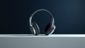 A single pair of sleek over-ear headphones resting on a minimal white surface against a deep navy background. Soft directional studio lighting catches the smooth curves of the ear cups and the glossy headband, casting a gentle shadow beneath. The headphones face slightly to the side, highlighting their modern sculptural form. Clean, sharp, and elegantly composed.
