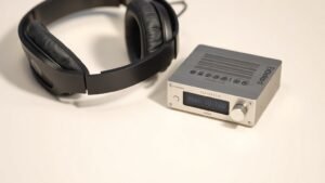 A high-end pair of over-ear headphones resting beside a compact desktop DAC and headphone amplifier unit on a clean off-white surface. Soft directional side lighting reveals the texture of the ear pads and the brushed aluminium of the DAC chassis. Photorealistic product photography, 16 by 9 horizontal format, no people, no text.