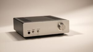 A compact headphone amplifier sitting on a clean off-white studio surface, photographed from a low front angle with soft side lighting revealing brushed aluminium fascia and front panel controls, deep shadow to the right, no background clutter, photorealistic product photography in 16 to 9 horizontal format